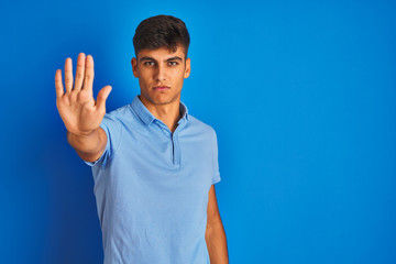 Young indian man wearing casual polo standing over isolated blue background doing stop sing with...