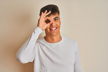 Young indian man wearing sweater standing over isolated white background doing ok gesture with hand smiling, eye looking through fingers with happy face.