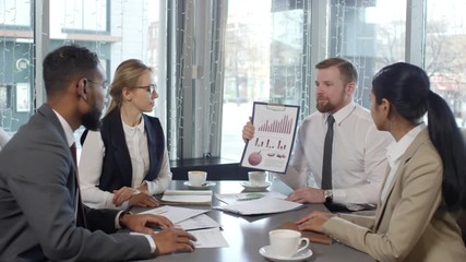 Zoom in of multi ethnic business team sitting at cafe table beside the window and discussing financial report during coffee break: Caucasian man explaining financial diagrams on clipboard - Powered by Adobe