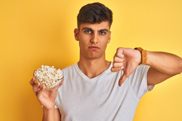 Young indian man holding pack of popcorn standing over isolated yellow background with angry face, negative sign showing dislike with thumbs down, rejection concept