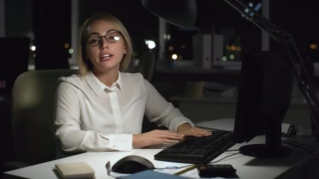 Medium shot of blond businesswoman in glasses finishing work on computer in office late in the evening. She is typing on keyboard, then turning towards camera and ranting about working overtime