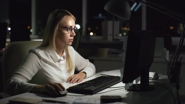 Medium shot of stressed businesswoman in glasses working on computer in dark office after hours. She is getting angry and hitting keyboard in frustration, then throwing out sticky notes