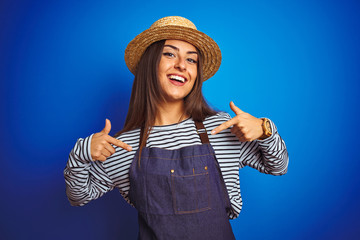 Young beautiful baker woman wearing apron and hat standing over isolated blue background looking...