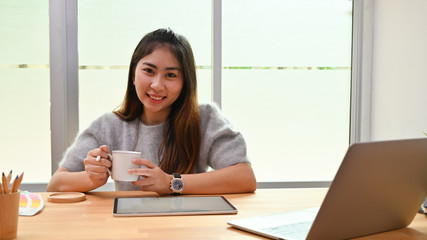 Young asian creative woman looking at camera and holding cup of coffee.