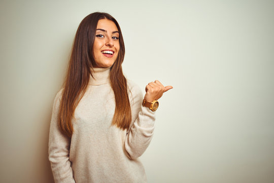 Young beautiful woman wearing winter turtleneck sweater over isolated white background smiling with happy face looking and pointing to the side with thumb up.