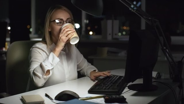 Medium shot of busy businesswoman in glasses working overtime in dark office. She is typing on computer and drinking coffee