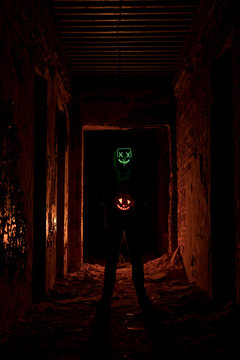 Man wearing glowing green scary mask standing in dark passage holding halloween pumpkin