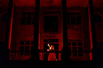 Girl in vintage dress standing on the balcony of old mansion with old lantern and halloween pumpkin lit by red light