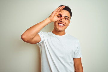 Fototapeta na wymiar Young brazilian man wearing t-shirt standing over isolated white background doing ok gesture with hand smiling, eye looking through fingers with happy face.