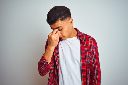 Young brazilian man wearing red shirt standing over isolated white background tired rubbing nose and eyes feeling fatigue and headache. Stress and frustration concept.
