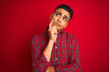 Young brazilian man wearing shirt standing over isolated red background with hand on chin thinking about question, pensive expression. Smiling with thoughtful face. Doubt concept.