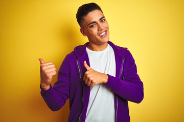 Young brazilian man wearing purple sweatshirt standing over isolated yellow background Pointing to the back behind with hand and thumbs up, smiling confident