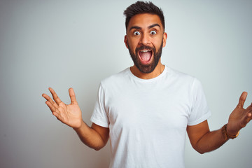 Young indian man wearing t-shirt standing over isolated white background celebrating crazy and amazed for success with arms raised and open eyes screaming excited. Winner concept