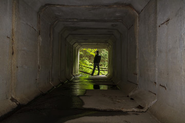 Young man standing on end of concrete stream tunnel in black jacket with backpack