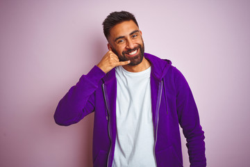 Young indian man wearing purple sweatshirt standing over isolated pink background smiling doing phone gesture with hand and fingers like talking on the telephone. Communicating concepts.