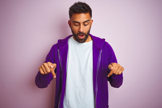 Young indian man wearing purple sweatshirt standing over isolated pink background Pointing down with fingers showing advertisement, surprised face and open mouth