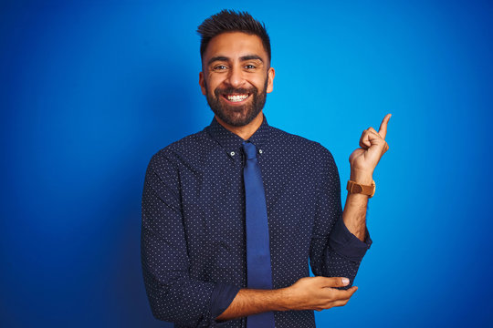 Young indian businessman wearing elegant shirt and tie standing over isolated blue background with a big smile on face, pointing with hand and finger to the side looking at the camera.