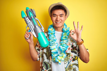 Asian chinese man wearing floral hawaian lei and water gun over isolated yellow background doing ok...