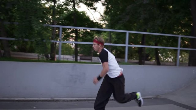 Sportive man practicing parkour in the open skate park - sliding from the ramp, crossing the barries amd making somersault in slow motion. Summer day, green trees around. Side view