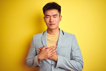 Young asian chinese businessman wearing jacket standing over isolated yellow background smiling with hands on chest with closed eyes and grateful gesture on face. Health concept.