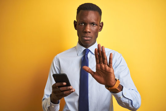 African american businessman using smartphone standing over isolated yellow background with open hand doing stop sign with serious and confident expression, defense gesture