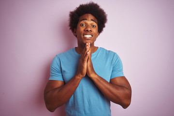 Obraz na płótnie Canvas African american man with afro hair wearing blue t-shirt standing over isolated pink background praying with hands together asking for forgiveness smiling confident.