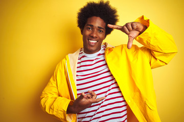 African american man with afro hair wearing rain coat standing over isolated yellow background gesturing with hands showing big and large size sign, measure symbol. Smiling looking at the camera