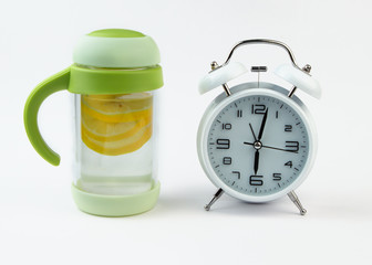 Alarm clock and a detox glass of water with lemon on white backgrounf. Wake up early in the morning by the alarm bell.