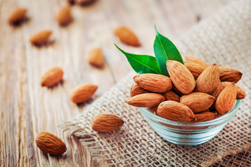 Almonds in glass bowl on rustic table with green leaves. Fresh almond on jute.