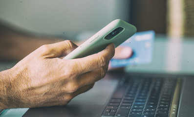 Man doing online shopping at home. Detail on hands holding smartphone and credit card with modern laptop keyboard on background