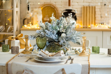 Beautiful served table with decorations, candles with glaze on white tablecloth on the background of the kitchen with people. Living room decorated with lights and Christmas tree.