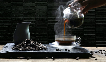 Man's hand pouring hot coffee into a glass coffee cup with steam on black stone plate and wooden table with black marble tiles wall background in vintage dark tone style 