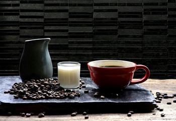 Selective focus at red coffee cup with milk and coffee beans on black slate and wooden table in black marble tile wall background