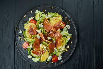 Close up view on Salad with honey pear, dor blue cheese and prosciutto on stylish dark plate on black wooden background. Selective focus
