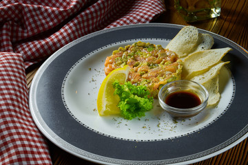 Fresh Salmon tartare, avocado with croutons and soy sauce in white plate on wooden background in composition with red cloth. Copy space. Top view