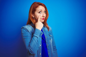 Young beautiful redhead woman wearing denim shirt standing over blue isolated background Pointing to the eye watching you gesture, suspicious expression