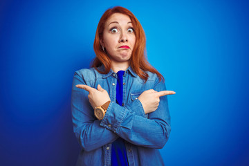 Fototapeta premium Young beautiful redhead woman wearing denim shirt standing over blue isolated background Pointing to both sides with fingers, different direction disagree