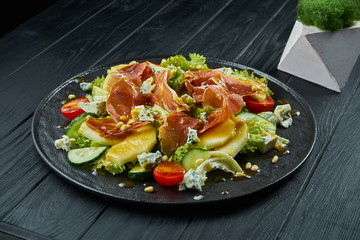 Close up view on Salad with honey pear, dor blue cheese and prosciutto on stylish dark plate on black wooden background. Selective focus