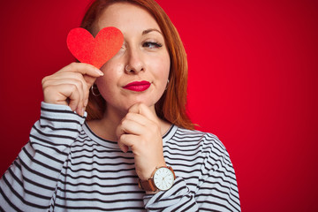 Young redhead romantic woman holding heart over red isolated background serious face thinking about question, very confused idea