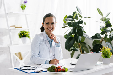 happy nutritionist in white coat near vegetables and laptop - obrazy, fototapety, plakaty