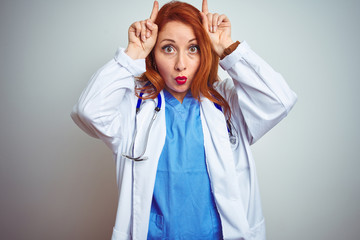 Young redhead doctor woman using stethoscope over white isolated background doing funny gesture with finger over head as bull horns