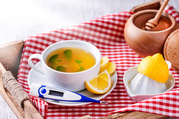 Thermometer and cup of tea with lemon on wooden tray. Honey in background with honey stick.