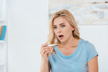 shocked woman holding digital thermometer at home