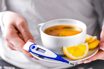Cup of tea and digital thermometer in woman hands. Pills, ginger.and lemon for illness prevention.