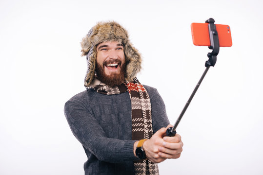 Cheerful bearded man is making a photo with selfie stick. He is wearing winter accessories.