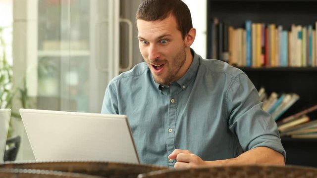Excited man using a laptop celebrating good news sitting in a coffee shop