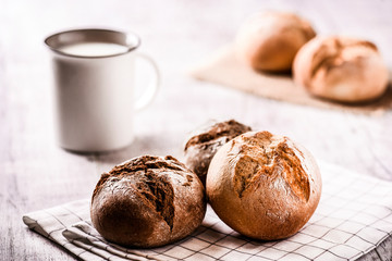Fresh hot buns and jar of milk in background. Dark rolls of tasty and  beautiful dark bread on white table.