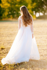 Fototapeta na wymiar Beautiful, romantic blonde girl dressed in a wedding dress is walking on a field, looking back in the sunlight.