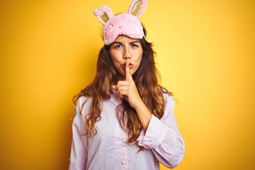 Young woman wearing pajama and sleep mask standing over yellow isolated background asking to be quiet with finger on lips. Silence and secret concept.