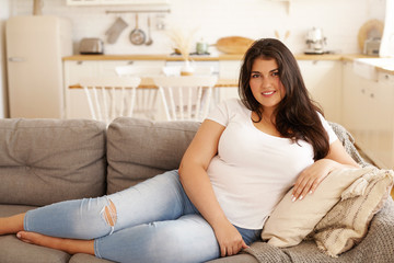 People, lifestyle, rest and relaxation concept. Attractive happy young Latin woman in casual clothes relaxing indoors after hard working day, stretching bare feet on sofa, smiling, being in good mood